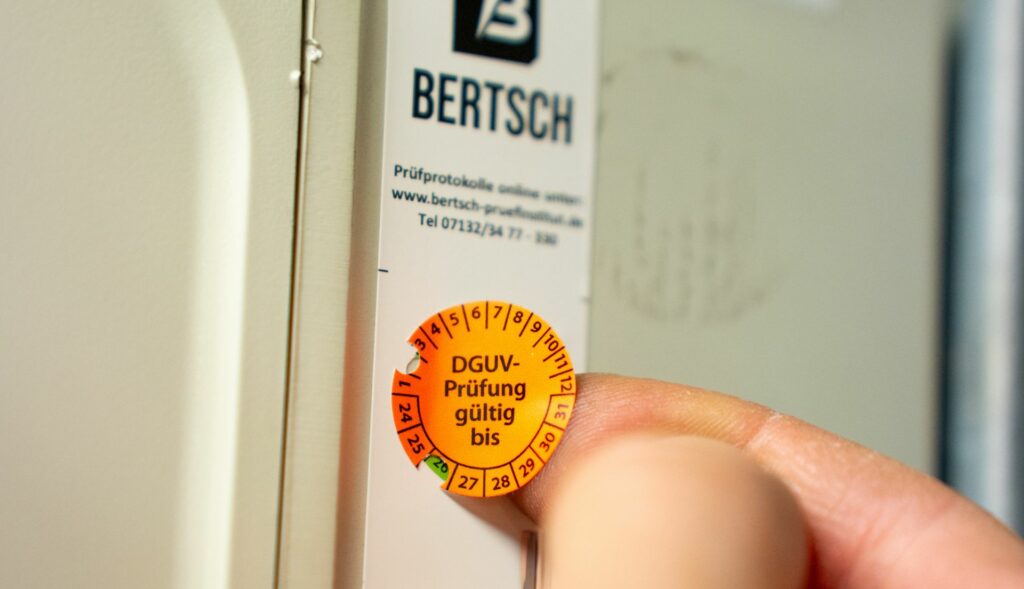 Eine Prüfplakette wird auf ein elektrisches Gerät geklebt mit der Info, dass die DGUV-Prüfung gültig ist bis Februar 2026